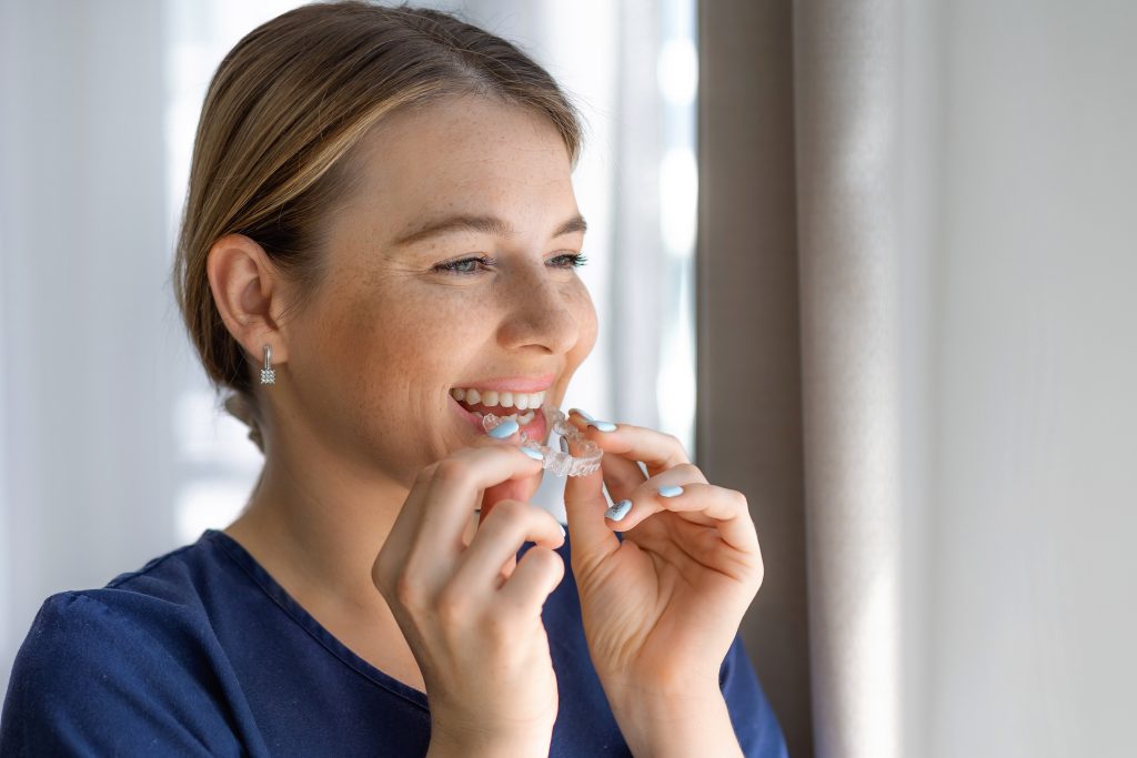 A woman holds and prepares to use a transparent dental aligner, smiling joyfully indoors.